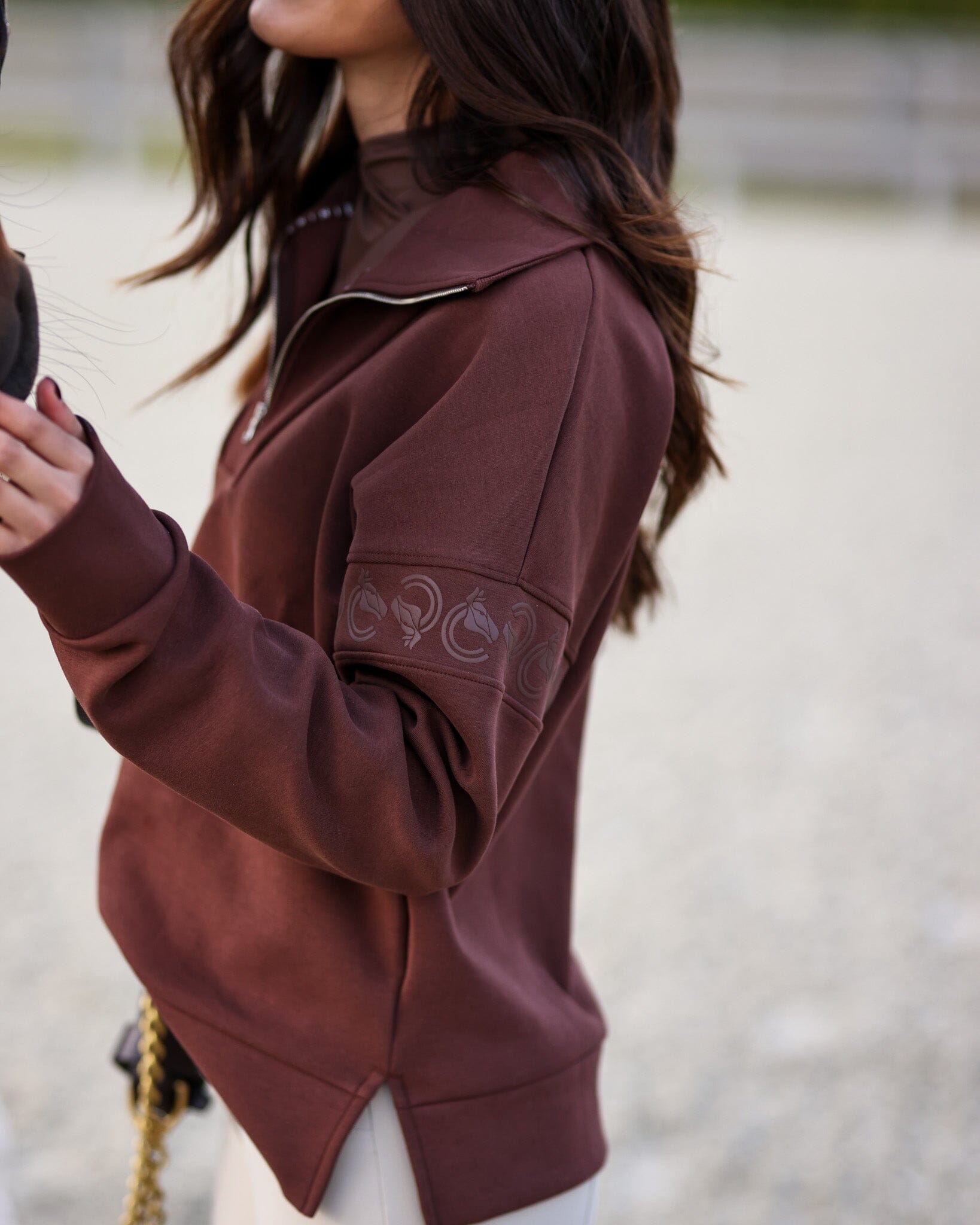 Side view of a female model wearing the Mocha Criniere Claire Sweater while standing next to a horse in an outdoor arena