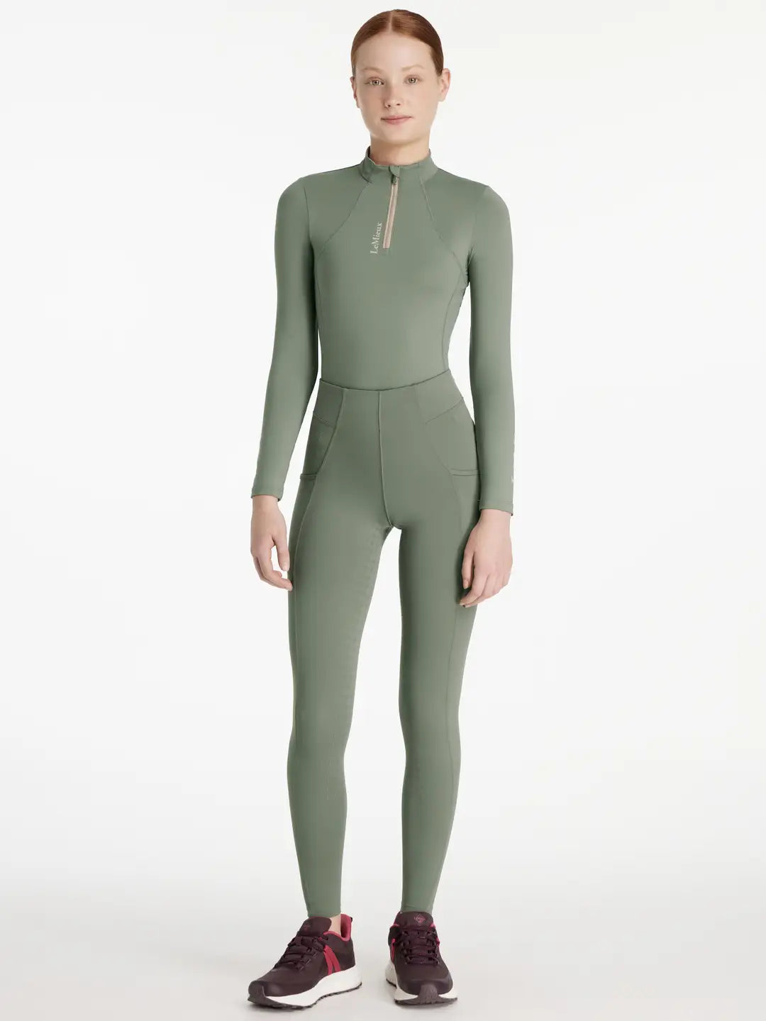 Full body front view of a young female model wearing the green Lemieux Young Rider Orla Pull On Tight and coordinating top on a white background