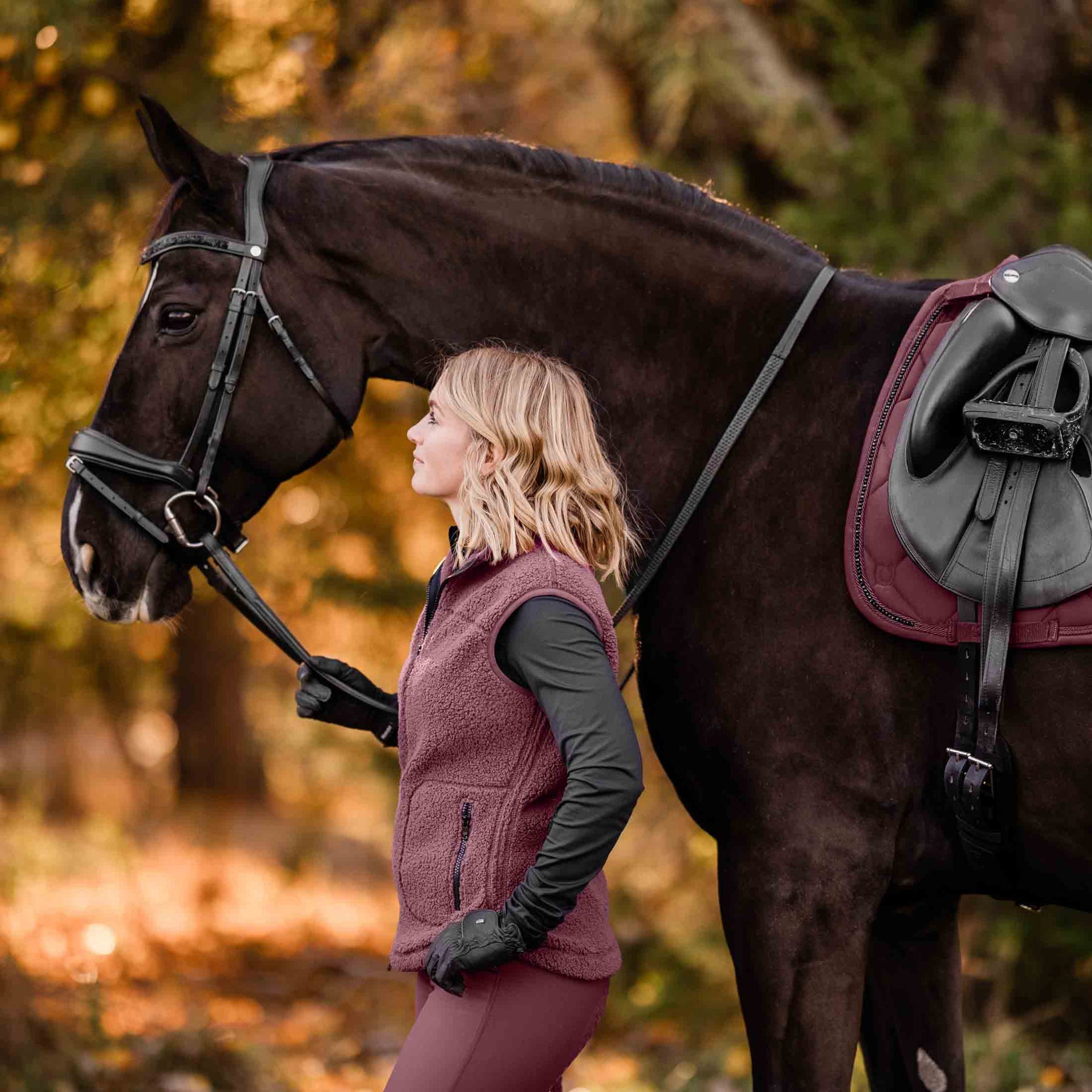 Side view of a horse wearing the Burgundy Equinavia Bergen Crystal Dressage Saddle Pad under a black saddle with a female model standing beside the horse