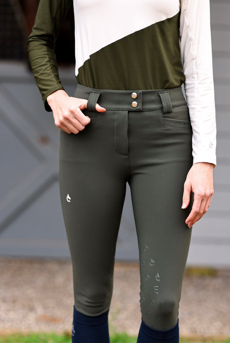 Front view of a female model wearing the Hunter Green Criniere Juliette High Compression Half Seat Breeches while standing in front of a barn