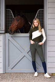 Front view of a female model wearing the Hunter Green Criniere Juliette High Compression Half Seat Breeches while standing in front of a horse in a stall