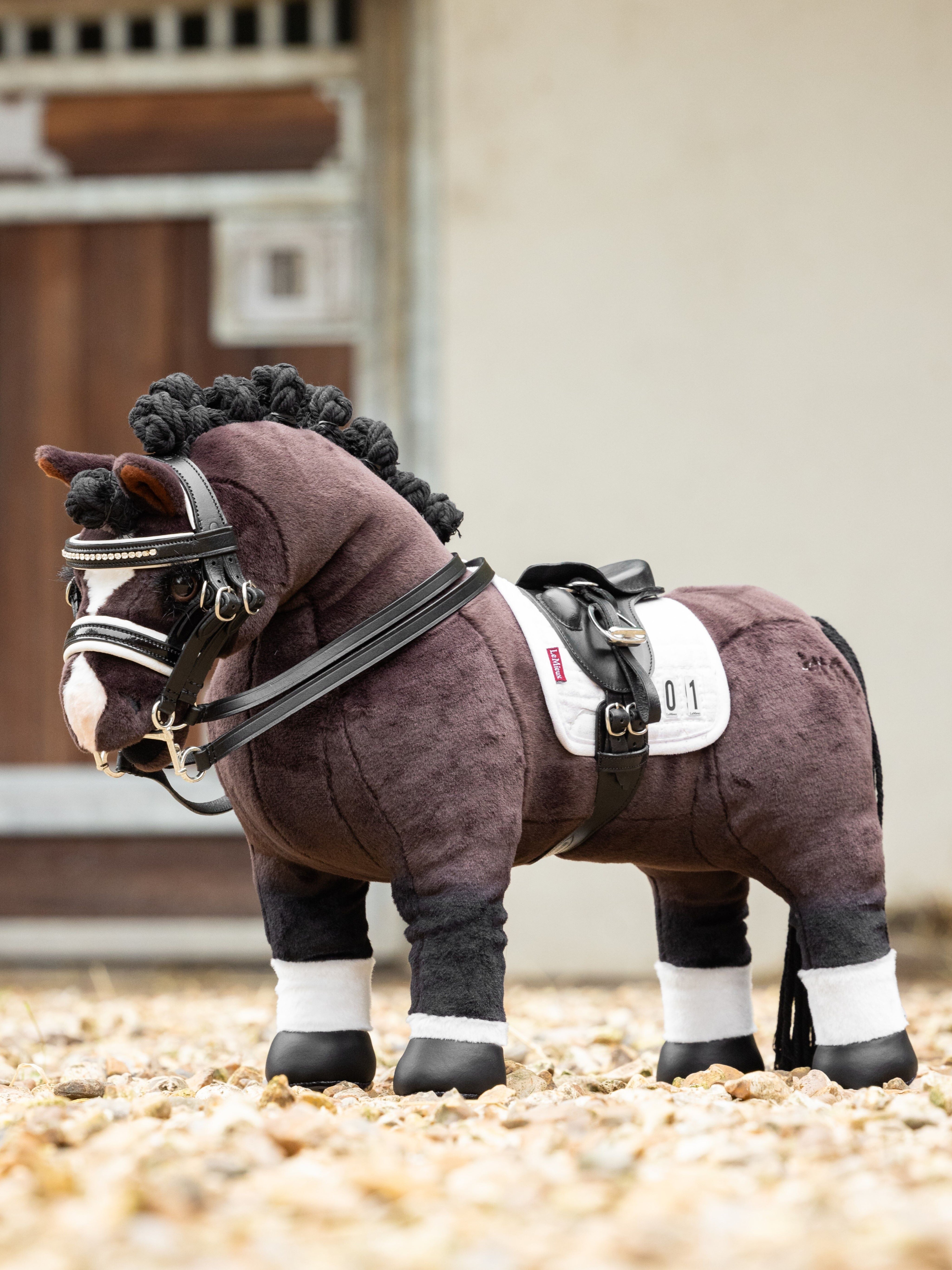 Side view of a bay toy pony wearing the LeMieux Toy Pony Dressage Saddle and Girth while standing on a gravel path