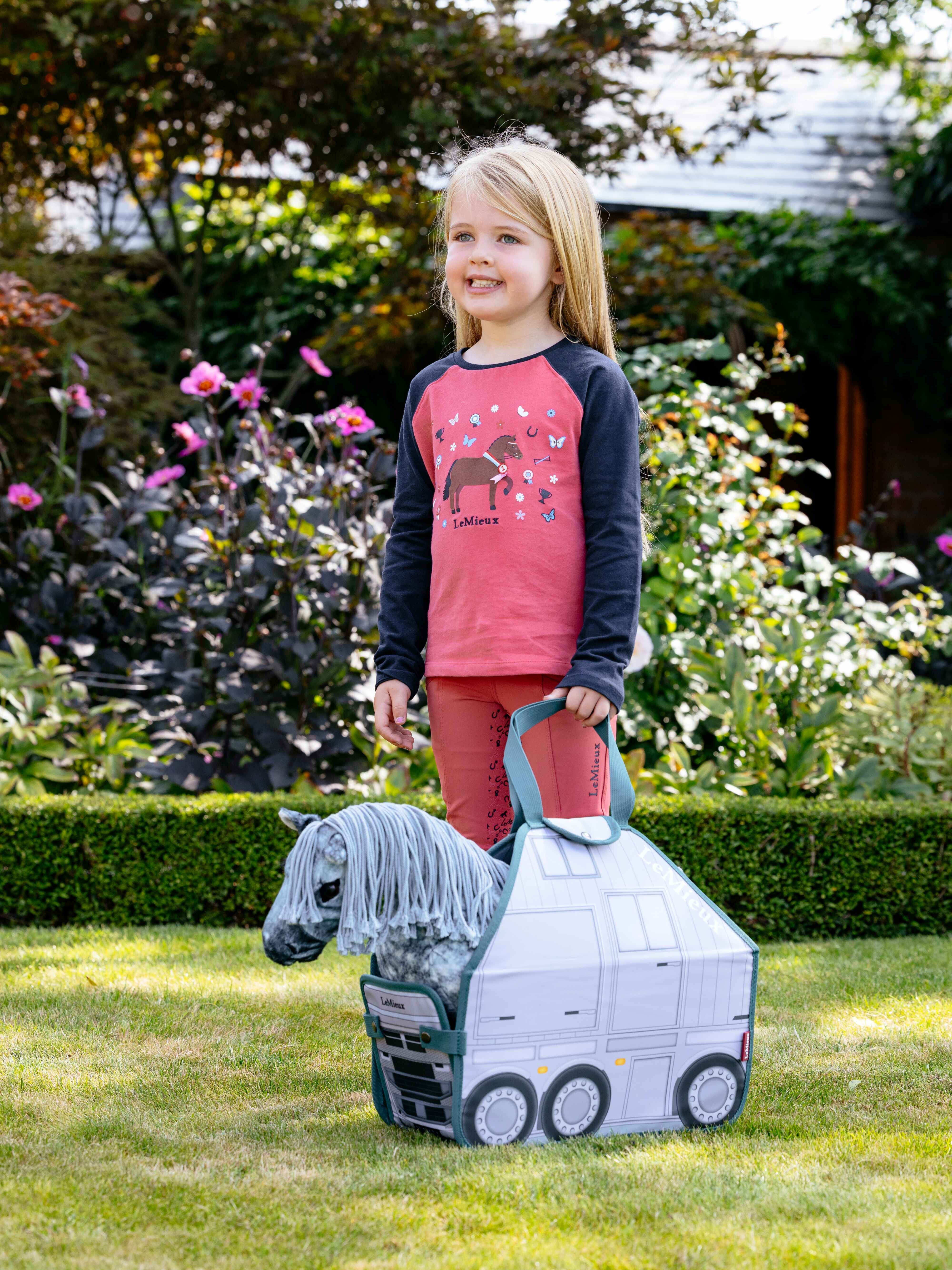 View of a young female model posing with a gray toy pony in her LeMieux Toy Pony Carry Bag while standing in the grass outside
