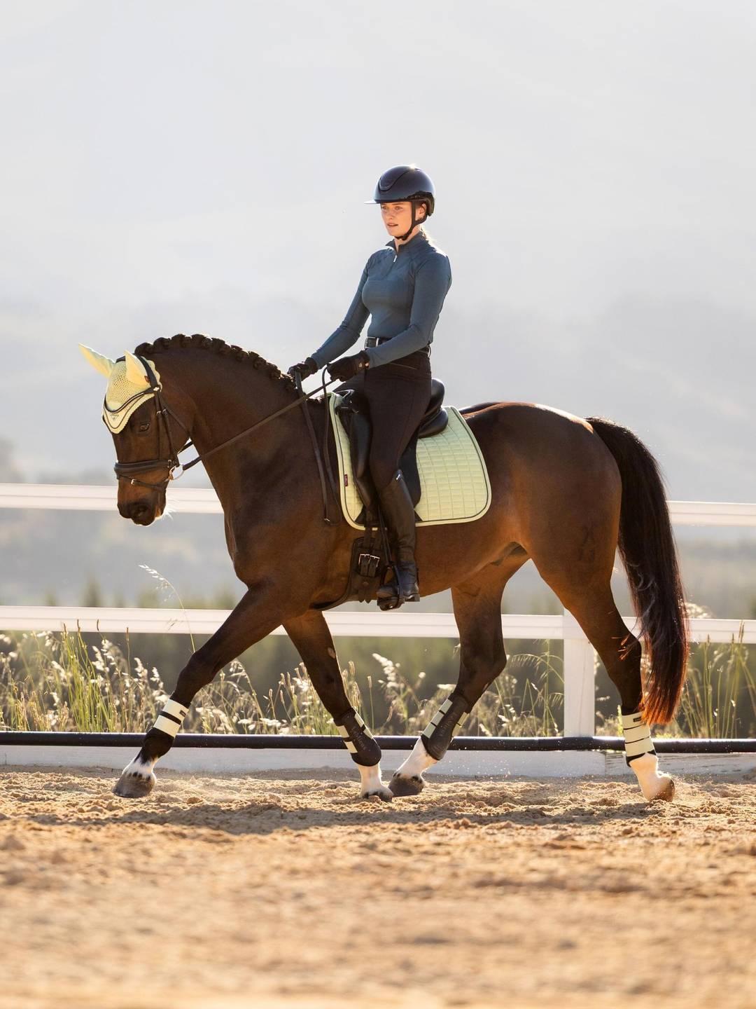 Side view of a model riding a horse that is wearing the macaron LeMieux Suede Dressage Square Pad