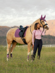 Front view of a model standing in front of a palomino horse wearing the mallow LeMieux Suede Dressage Square Pad and matching accessories while standing in a pasture
