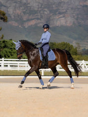Side view of a model riding a horse that is wearing the powder blue LeMieux Suede Dressage Square Pad