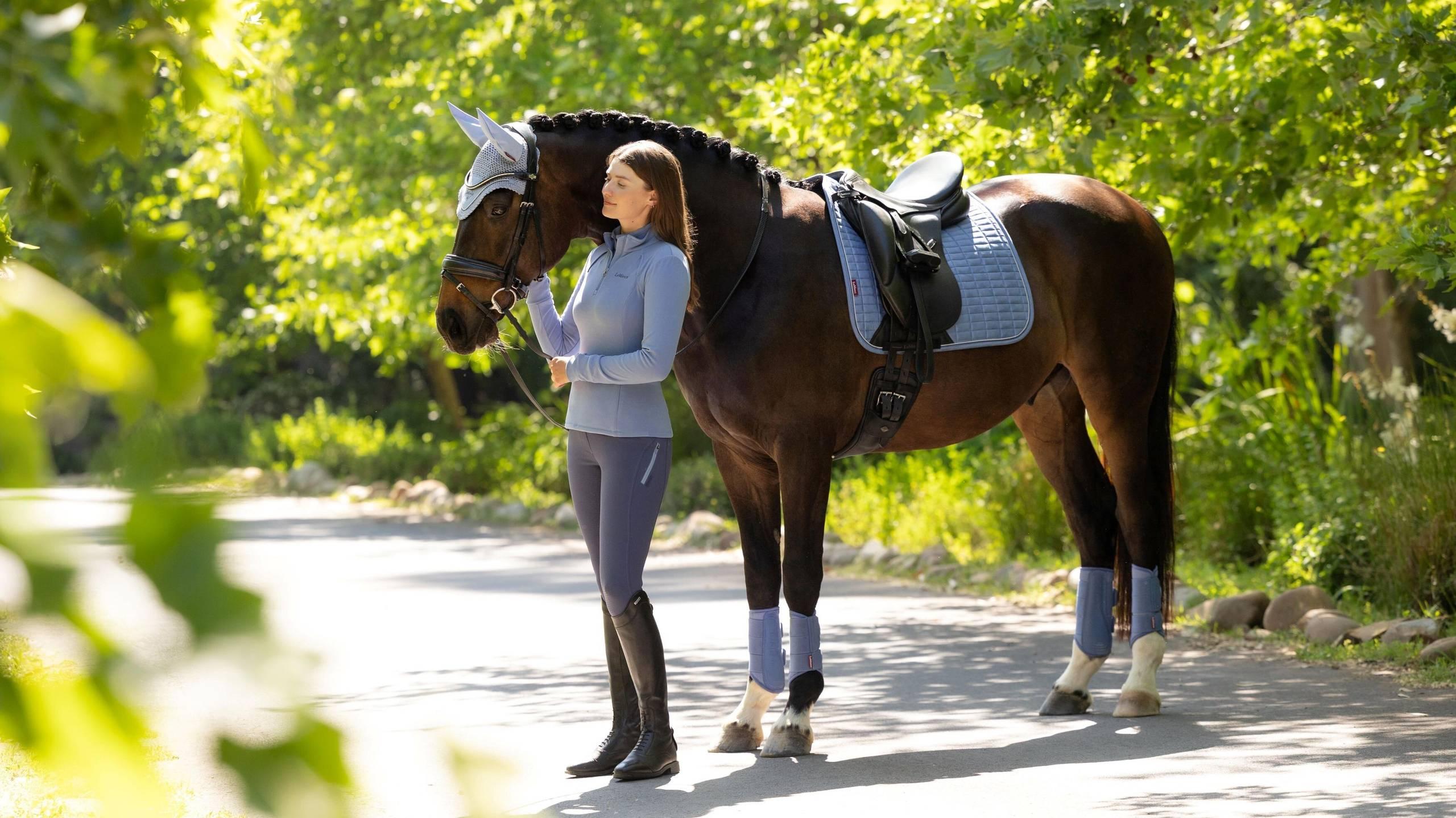 Side view of a model standing in front of a dark colored horse that is wearing the powder blue LeMieux Suede Dressage Square Pad and matching accessories