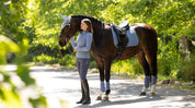Side view of a model standing in front of a dark colored horse that is wearing the powder blue LeMieux Suede Dressage Square Pad and matching accessories