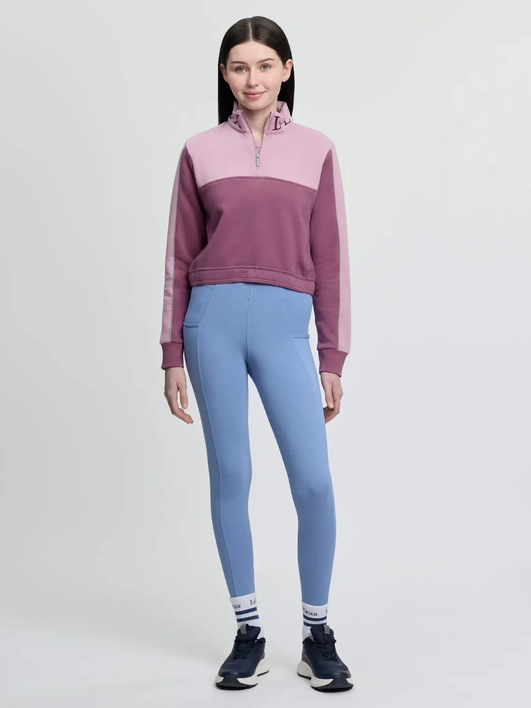 Full body front view of a young female model wearing the pink LeMieux Young Rider Cropped Kate Quarter Zip and blue tights on a plain background