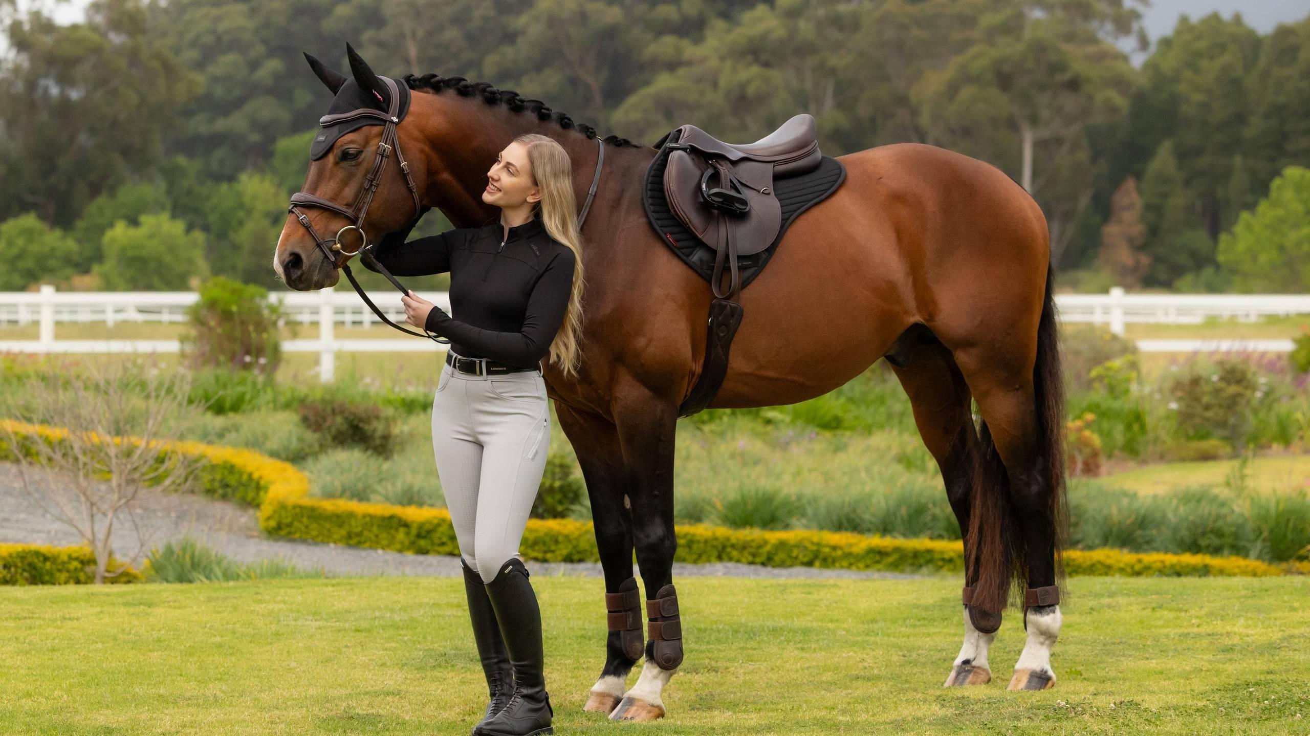 Side view of a female model standing in front of a bay horse that is wearing the black LeMieux Air-Tek Mesh Half Square Pad