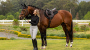 Side view of a female model standing in front of a bay horse that is wearing the black LeMieux Air-Tek Mesh Half Square Pad