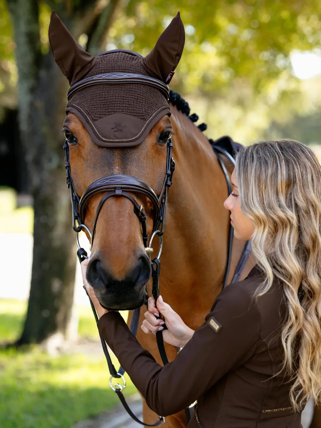 Front view of a bay horse wearing the brown LeMieux Brilliance Fly Hood while standing next to a female model
