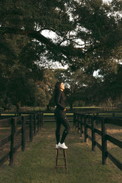 Side view of a female model wearing the Black Criniere Juliette High Compression Half Seat Breeches while standing on a stool outside