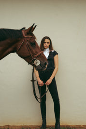 Front view of a female model wearing the Black Criniere Juliette High Compression Half Seat Breeches while standing behind a horse's head