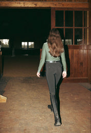 Back view of a female model wearing the Black Criniere Juliette High Compression Half Seat Breeches while walking into an indoor arena