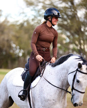 Front view of a female model wearing the Mocha Criniere Juliette High Compression Half Seat Breeches and coordinating top while riding a gray horse outside