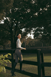 Side view of a female model wearing the Perfect Gray Criniere Juliette High Compression Half Seat Breeches while standing on the lower panel of a black fence outside