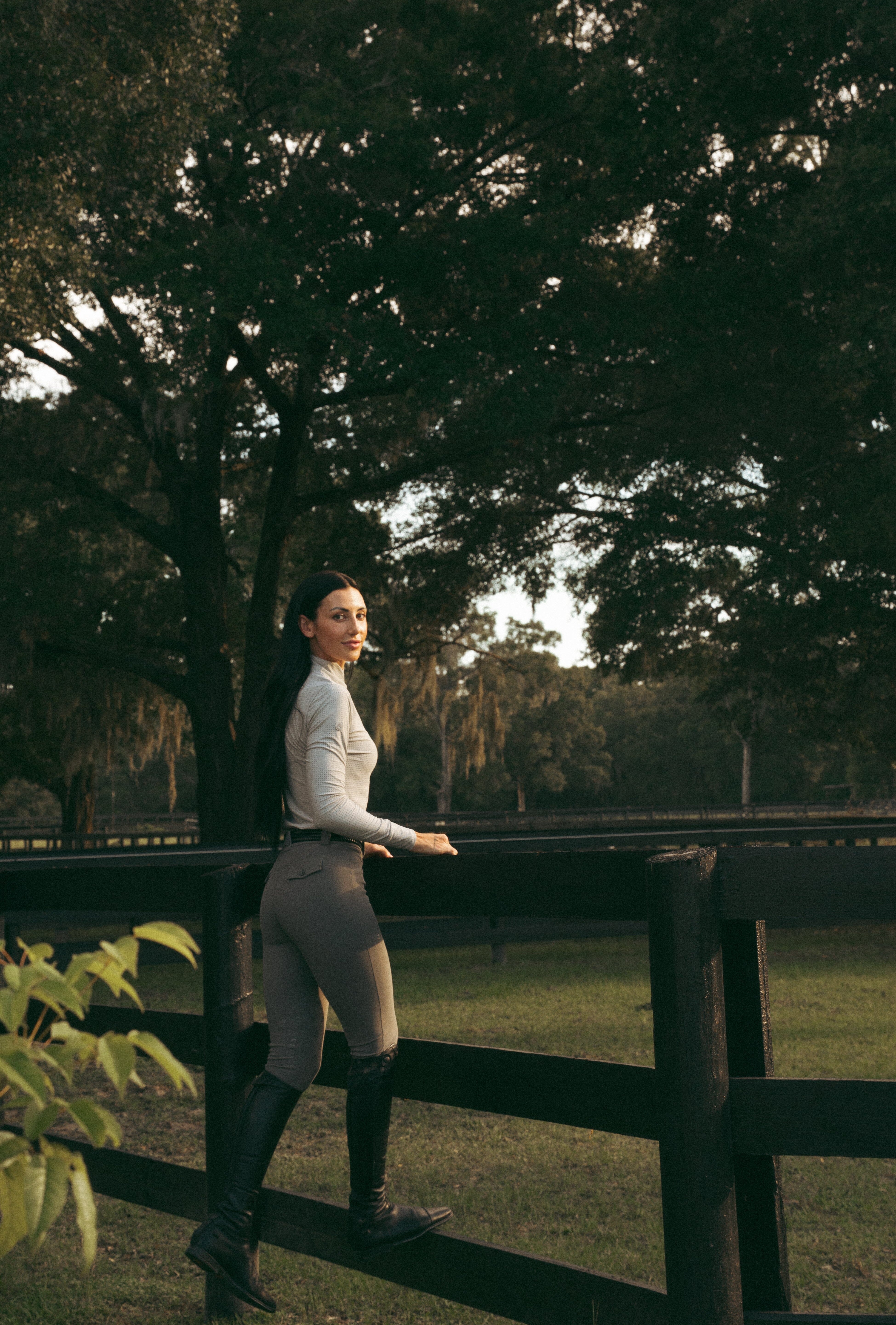 Side view of a female model wearing the Perfect Gray Criniere Juliette High Compression Half Seat Breeches while standing on the lower panel of a black fence outside