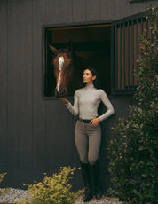 Full body front view of a female model wearing the Perfect Gray Criniere Juliette High Compression Half Seat Breeches and coordinating top while standing in front of a horse that is in a stall