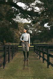 Full body front view of a female model wearing the Perfect Gray Criniere Juliette High Compression Half Seat Breeches and coordinating top while standing on a stool outside