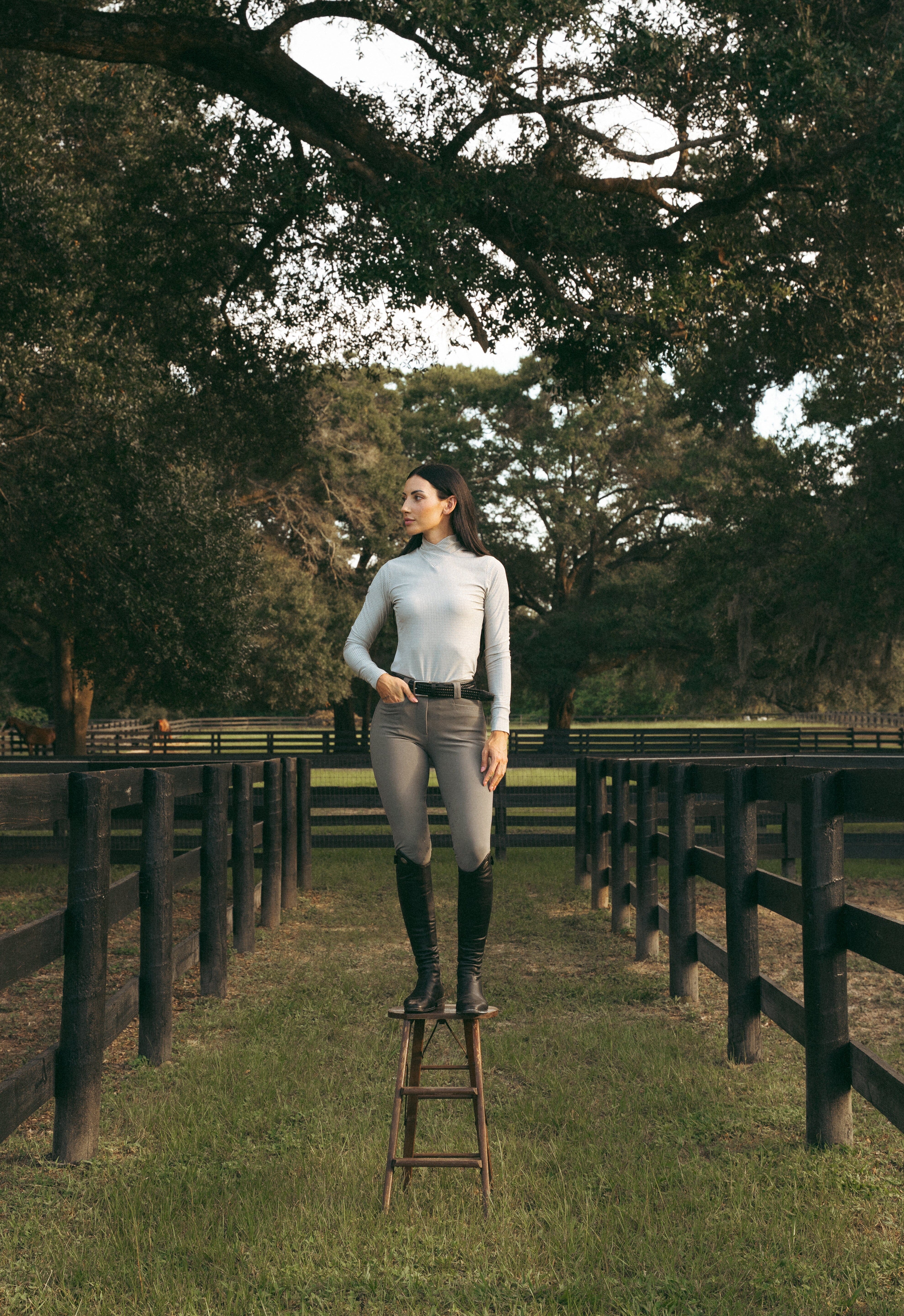 Full body front view of a female model wearing the Perfect Gray Criniere Juliette High Compression Half Seat Breeches and coordinating top while standing on a stool outside