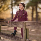 Front view of a young model wearing the Burgundy Equinavia Lena Kids Long Sleeve Quarter Zip outdoors standing up against a fence