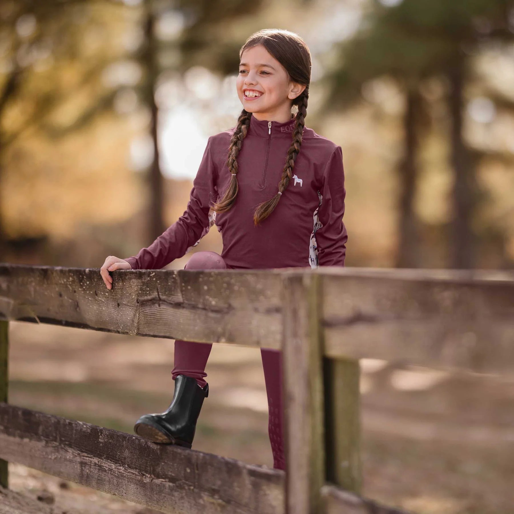 Front view of a young model wearing the Burgundy Equinavia Lena Kids Long Sleeve Quarter Zip outdoors standing up against a fence