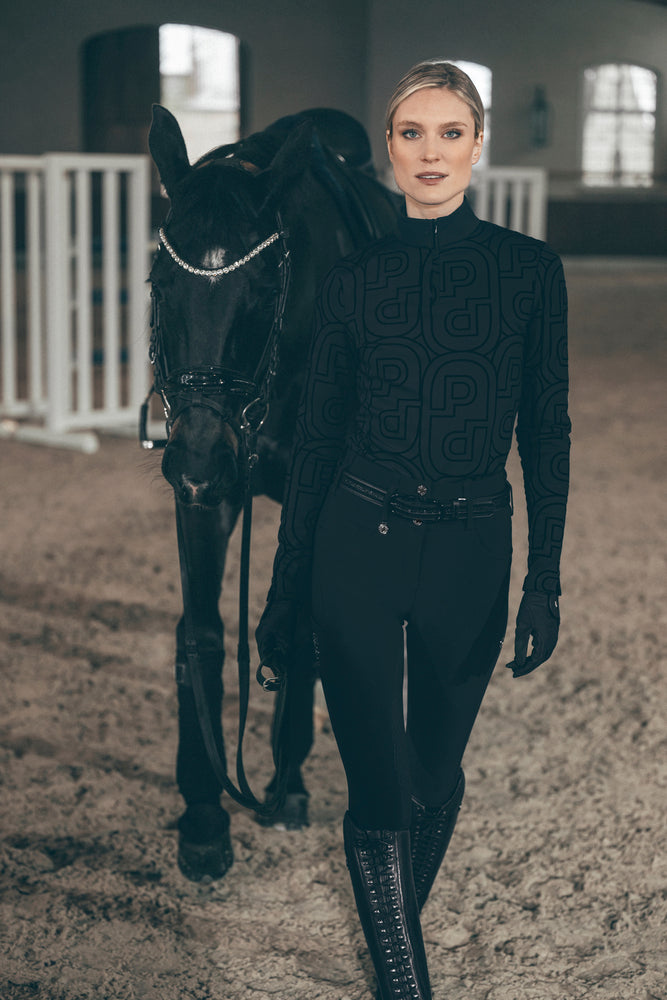 Front view of a female model wearing the Black Pikeur Zip Print Shirt while leading a black horse through an indoor arena