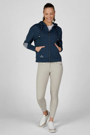 Full body front view of a female model wearing the Pine Green Pikeur Athleisure Zip Hoody with tan breeches on a plain background