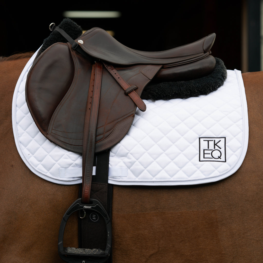 Side view of a horse wearing the White TKEQ x SPR Jump Saddle Pad under a brown jump saddle